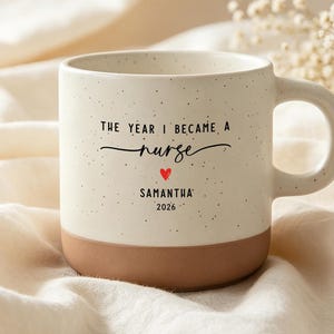 Op de afbeelding: Een gespikkelde, crèmekleurige keramische mok met een bruine onderkant. De tekst luidt: THE YEAR I BECAME A nurse, gevolgd door een klein rood hartje, de naam SAMANTHA en het jaartal 2026. De mok heeft een zijhandvat en een rond ontwerp.