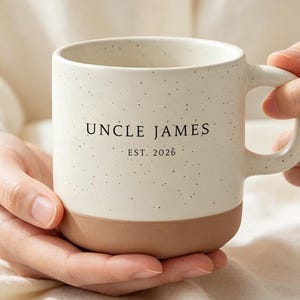 May include: A ceramic mug with a speckled cream-colored upper section and a solid terracotta-colored base. The text UNCLE JAMES EST. 2026 is printed in black on the front. The mug features a curved handle on the right side and a smooth finish.