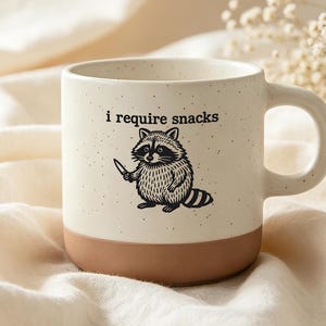 Op de afbeelding: Een crèmekleurige keramische mok met een gespikkelde afwerking en een effen terracotta-kleurige basis. De voorzijde toont een zwarte lijntekening van een wasbeer die een klein mes vasthoudt, met de tekst "i require snacks" boven de wasbeer.