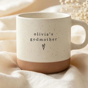 Könnte beinhalten: Ein gesprenkelter, cremefarbener Keramikbecher mit braunem Boden und Henkel. Der Text 'olivia's godmother' ist in Schwarz auf der Vorderseite aufgedruckt, ergänzt durch ein kleines, handgezeichnetes Herzsymbol unter dem Text.