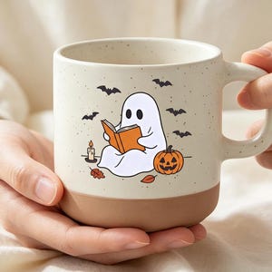 May include: A cream-colored ceramic mug with a speckled finish and a solid brown base. The front features a graphic of a white ghost reading an orange book, surrounded by a jack-o'-lantern, a lit candle, fallen leaves, and small black bats.