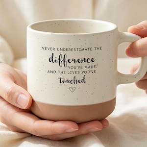 Może przedstawiać: Kremowy kubek ceramiczny z nakrapianym wykończeniem i jednolitym jasnobrązowym dnem. Z przodu znajduje się czarny napis: NEVER UNDERESTIMATE THE difference YOU'VE MADE, AND THE LIVES YOU'VE touched, oraz mała ikona serca.