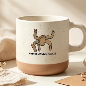 Pode incluir: Uma caneca de cerâmica branca suja com pintas e uma base sólida em tom castanho claro. A frente apresenta uma ilustração de linhas castanhas de uma criatura com várias pernas a fazer sinais de paz, com o texto preto "AMAZE AMAZE AMAZE" abaixo.