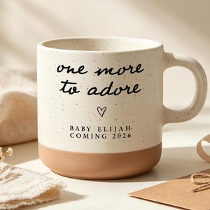 May include: A speckled cream-coloured ceramic mug with a terracotta-coloured base. The front features black text that reads one more to adore, a small heart symbol, and the text BABY ELIJAH COMING 2026 in a clean, sans-serif font.