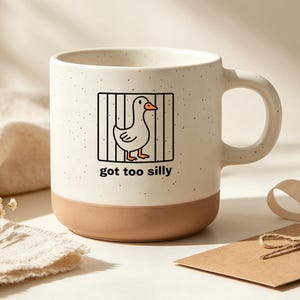 Pode incluir: Uma caneca de cerâmica branca suja com pintas, base lisa castanha clara e uma pega. A frente apresenta um desenho de linha preta de um pato dentro de uma gaiola quadrada com o texto "got too silly" impresso em letras minúsculas pretas.