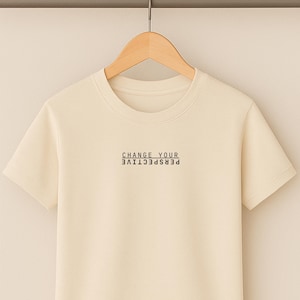 Puede incluir: Camiseta de manga corta color crema con el texto negro "CHANGE YOUR PERSPECTIVE". La camiseta está colgada de una percha de madera sobre un fondo neutro.