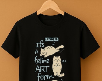 May include: Black t-shirt with a graphic of three cartoon cats in various poses, with the text "It's A feline ART form" and "LAZINESS:" above the cats. The cats are light brown with black outlines.