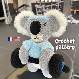Könnte beinhalten: Ein gehäkelter Koala mit blauem Stirnband und Hemd, der eine schwarze Langhantel hält. Der Koala ist grau mit weißem Fell um Kopf und Ohren. Der Text "Crochet pattern" ist sichtbar.