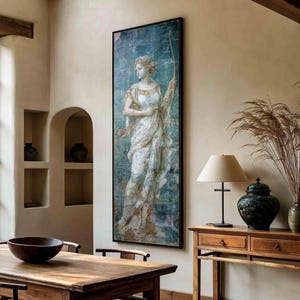 May include: A tall rectangular wall art print featuring a classical figure in a flowing white gown holding a bow, set against a textured blue background. The artwork is framed in a thin black border and displayed on a light-colored interior wall.