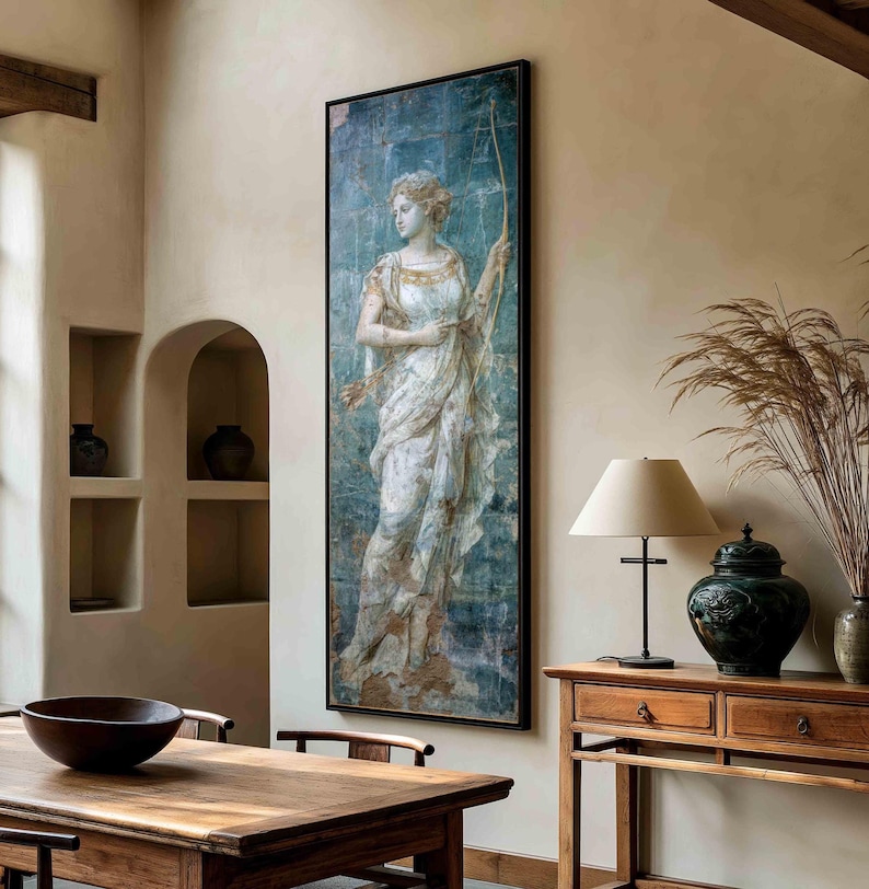 May include: A tall rectangular wall art print featuring a classical figure in a flowing white gown holding a bow, set against a textured blue background. The artwork is framed in a thin black border and displayed on a light-colored interior wall.