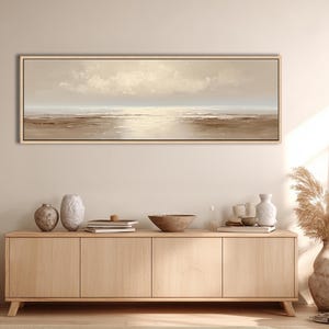 May include: A long, rectangular landscape painting with a neutral color palette. The artwork depicts a serene seascape with a light-filled sky and calm water. The painting is framed in a light wood tone and hangs above a wooden cabinet.