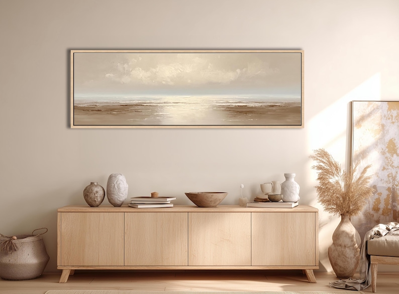 May include: A long, rectangular landscape painting with a neutral color palette. The artwork depicts a serene seascape with a light-filled sky and calm water. The painting is framed in a light wood tone and hangs above a wooden cabinet.
