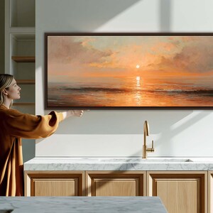 May include: A panoramic landscape painting in a dark brown frame depicting a sunset over the ocean. The artwork features soft orange and peach hues in the sky with clouds and a bright sun reflecting on the calm water surface below the horizon line.