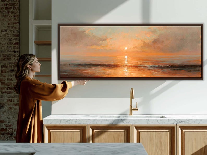 May include: A panoramic landscape painting in a dark brown frame depicting a sunset over the ocean. The artwork features soft orange and peach hues in the sky with clouds and a bright sun reflecting on the calm water surface below the horizon line.