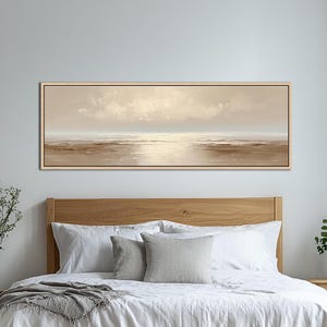 May include: A long, rectangular landscape painting in a light beige color palette. The artwork depicts a serene coastal scene with a soft, cloudy sky and calm water. The painting is framed in a light wood tone and hangs above a bed.