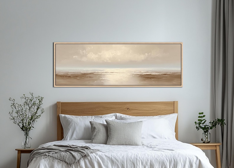 May include: A long, rectangular landscape painting in a light beige color palette. The artwork depicts a serene coastal scene with a soft, cloudy sky and calm water. The painting is framed in a light wood tone and hangs above a bed.