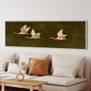 Peut inclure: Une œuvre d'art encadrée représentant trois grues blanches en vol sur un fond vert foncé. Les oiseaux sont représentés avec leurs ailes déployées, créant une sensation de mouvement. L'œuvre est exposée au-dessus d'un canapé avec des coussins de couleur neutre.