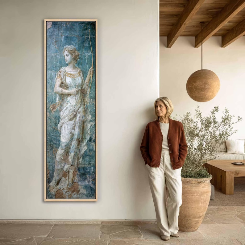 May include: A tall, rectangular framed wall art piece featuring a classical painting of a figure in a flowing white gown holding a long bow. The artwork has a distressed blue and gold background, displayed in a light wood frame on a white wall.