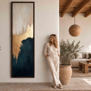 Puede incluir: Una pieza de arte mural vertical, alta y rectangular, enmarcada en madera oscura, que presenta una composición abstracta con una base azul marino profundo, una banda central de pan de oro metálico y una sección superior blanca.