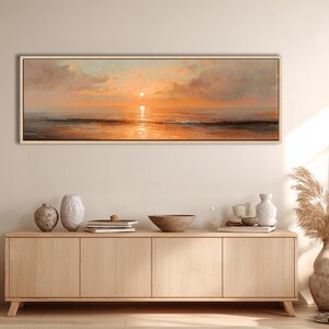 May include: A panoramic landscape painting in a light wood frame depicting a sunset over the ocean. The sky features soft clouds in shades of orange, pink, and pale blue, with the sun reflecting on the calm water surface below the horizon line.