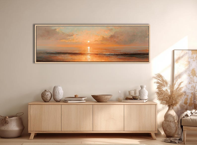 May include: A panoramic landscape painting in a light wood frame depicting a sunset over the ocean. The sky features soft clouds in shades of orange, pink, and pale blue, with the sun reflecting on the calm water surface below the horizon line.