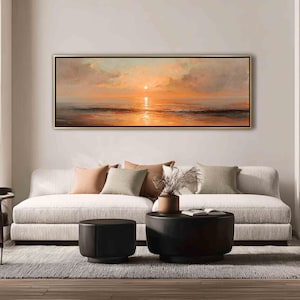 May include: A panoramic landscape painting in a thin gold frame depicting a sunset over the ocean. The scene features soft orange and peach hues in the sky and clouds, with a bright sun reflecting on the calm water surface below the horizon line.