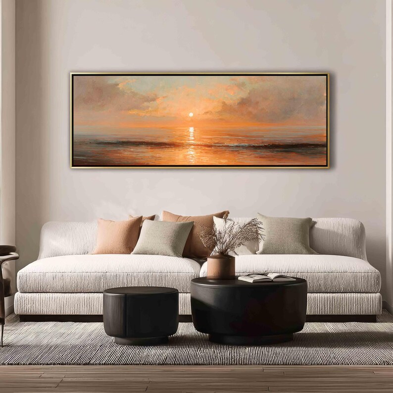 May include: A panoramic landscape painting in a thin gold frame depicting a sunset over the ocean. The scene features soft orange and peach hues in the sky and clouds, with a bright sun reflecting on the calm water surface below the horizon line.