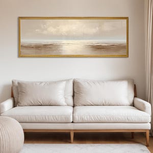 May include: A long, rectangular framed painting depicting a serene seascape in muted tones of beige, brown, and cream. The artwork hangs above a cream-colored sofa with wooden legs. The room is illuminated by natural light.