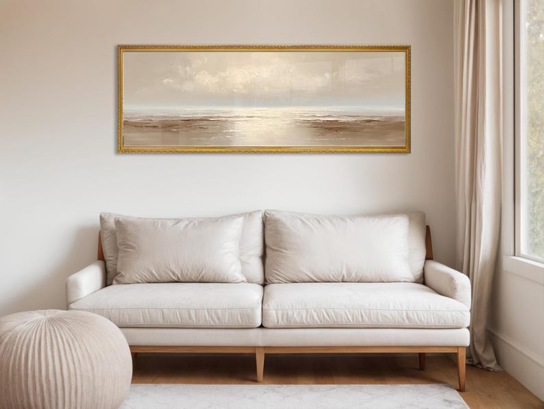 May include: A long, rectangular framed painting depicting a serene seascape in muted tones of beige, brown, and cream. The artwork hangs above a cream-colored sofa with wooden legs. The room is illuminated by natural light.