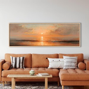 May include: A panoramic landscape painting in a light wood frame depicting a sunset over the ocean. The artwork features soft clouds in shades of orange, pink, and gold, with the sun reflecting on the calm water surface and dark waves below.