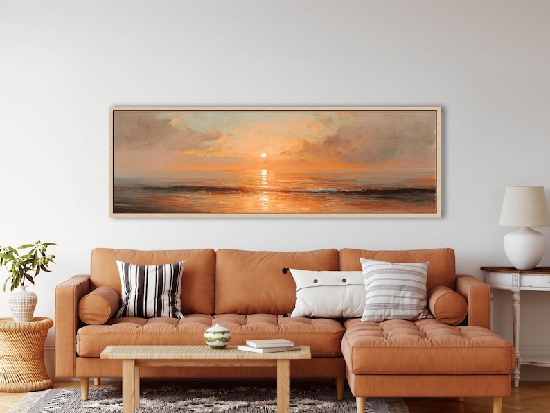 May include: A panoramic landscape painting in a light wood frame depicting a sunset over the ocean. The artwork features soft clouds in shades of orange, pink, and gold, with the sun reflecting on the calm water surface and dark waves below.