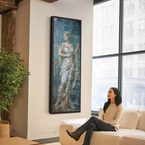 May include: A tall, rectangular framed wall art piece featuring a classical painting of a figure in a flowing white and gold garment holding a bow. The artwork has a distressed, weathered texture with blue and earthy brown tones in a dark frame.