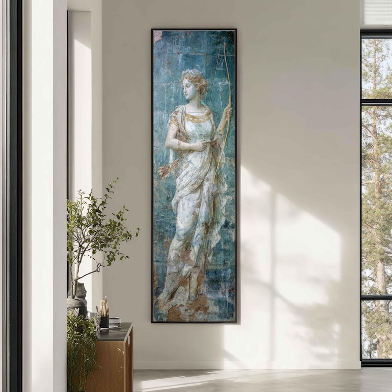 May include: A tall, rectangular wall art piece featuring a classical figure in a flowing white gown against a distressed, textured blue background. The figure holds a long, thin bow, with a quiver of arrows visible at the hip, framed in a thin black border.