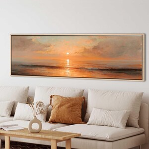 May include: A panoramic landscape painting in a light wood frame depicting a sunset over the ocean. The artwork features soft orange, gold, and peach hues reflecting on the water surface, with textured clouds and a calm horizon line.