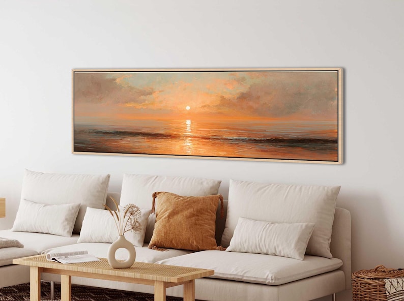May include: A panoramic landscape painting in a light wood frame depicting a sunset over the ocean. The artwork features soft orange, gold, and peach hues reflecting on the water surface, with textured clouds and a calm horizon line.