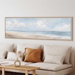 Puede incluir: Una pintura de paisaje panorámico en un marco de madera clara que representa una serena escena costera con una playa de arena, suaves olas azules del océano y un cielo nublado. La obra presenta tonos apagados de beige, blanco y azul claro.