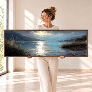 May include: A panoramic landscape painting in a dark wood frame depicting a serene lake at sunset. The scene features soft blue and golden light reflecting on the water, with tall grasses along the shoreline and distant mountains under a cloudy sky.