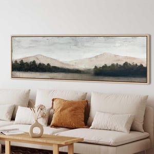 Puede incluir: Una pintura de paisaje panorámico en un marco de madera clara que presenta montañas de color beige suave bajo un cielo gris pálido, con una línea de bosque verde oscuro que se extiende por el primer plano. La obra muestra una paleta de colores.