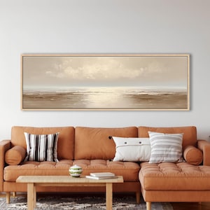 May include: A long, rectangular landscape painting with a light wood frame. The artwork depicts a serene coastal scene with a muted color palette of beige, brown, and white. The painting hangs above a rust-colored sectional sofa with decorative pillows.