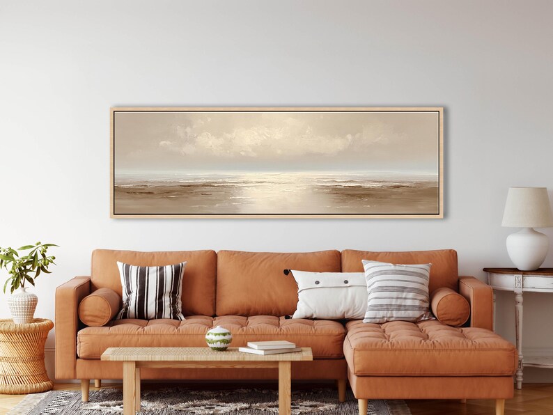 May include: A long, rectangular landscape painting with a light wood frame. The artwork depicts a serene coastal scene with a muted color palette of beige, brown, and white. The painting hangs above a rust-colored sectional sofa with decorative pillows.