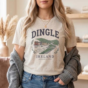 May include: A light beige t-shirt with the word "DINGLE" in navy blue arched across the top. Below the word is a green and brown illustration of a landscape with buildings and water. The word "IRELAND" is below the illustration.
