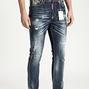 May include: Dark wash denim jeans with distressed details, including patches and paint splatters. The jeans have a classic five-pocket design and a straight leg fit. A white tag hangs from the front. The model is wearing black trainers.