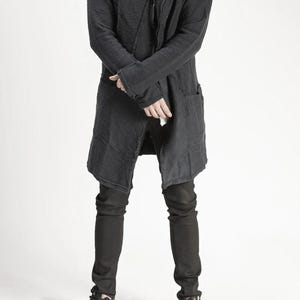 Könnte beinhalten: Ein dunkelgrauer langer Mantel mit strukturierter Optik, mit Reverskragen und Taschen. Der Mantel wird mit schwarzen Jeans und schwarzen Sneakers kombiniert. Der Gesamtstil ist modern und lässig.