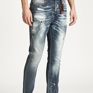 Könnte beinhalten: Blaue Jeans aus Denim mit Used-Waschung, Farbspritzern und einem verblichenen Effekt. Die Jeans haben ein klassisches Fünf-Taschen-Design und eine gerade Beinpassform. Ein schwarzes Etikett hängt an der Gürtelschlaufe. Die Person trägt schwarze Sneaker.