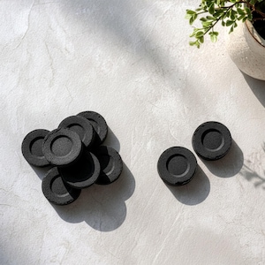 May include: A collection of small, circular black charcoal discs, each featuring a recessed centre. The discs are arranged in a cluster of several stacked pieces on the left side, with two additional individual discs positioned to the right.