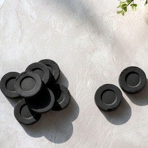 May include: A collection of circular black charcoal discs, each featuring a recessed centre. The discs are arranged in a cluster of six on the left side, with two additional individual discs positioned separately to the right.
