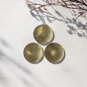 Op de afbeelding: Drie goudkleurige, cirkelvormige metalen onderzetters met een geperforeerd stippenpatroon. De onderzetters zijn in een driehoekige formatie op een wit oppervlak gerangschikt, met een takje witte bloemen en schaduwen op de achtergrond.