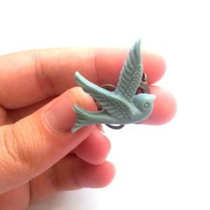 Puede incluir: Anillo de pájaro azul claro con alas extendidas. El pájaro está hecho de un material liso y está montado sobre una base de anillo de color bronce. El anillo es un accesorio decorativo.