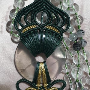 May include: A green and gold macrame pendant featuring a clear quartz stone. The pendant is suspended from a strand of clear quartz beads.