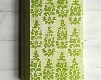 Op de afbeelding: Een hardcover notitieboek met een groene rug en een patroon op de kaft. De kaft heeft een herhalend ontwerp van groene bloemvormen op een crèmekleurige achtergrond. Het notitieboek is gesloten en ligt op een wit houten oppervlak.